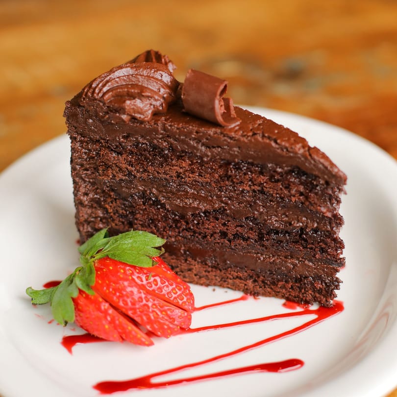 A slice of rich chocolate cake with frosting, chocolate curls, and a fresh strawberry on a white plate.