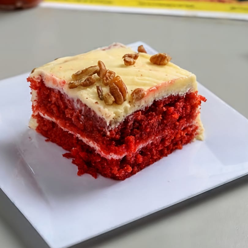 Slice of red velvet cake with cream cheese frosting and pecans on a white plate.