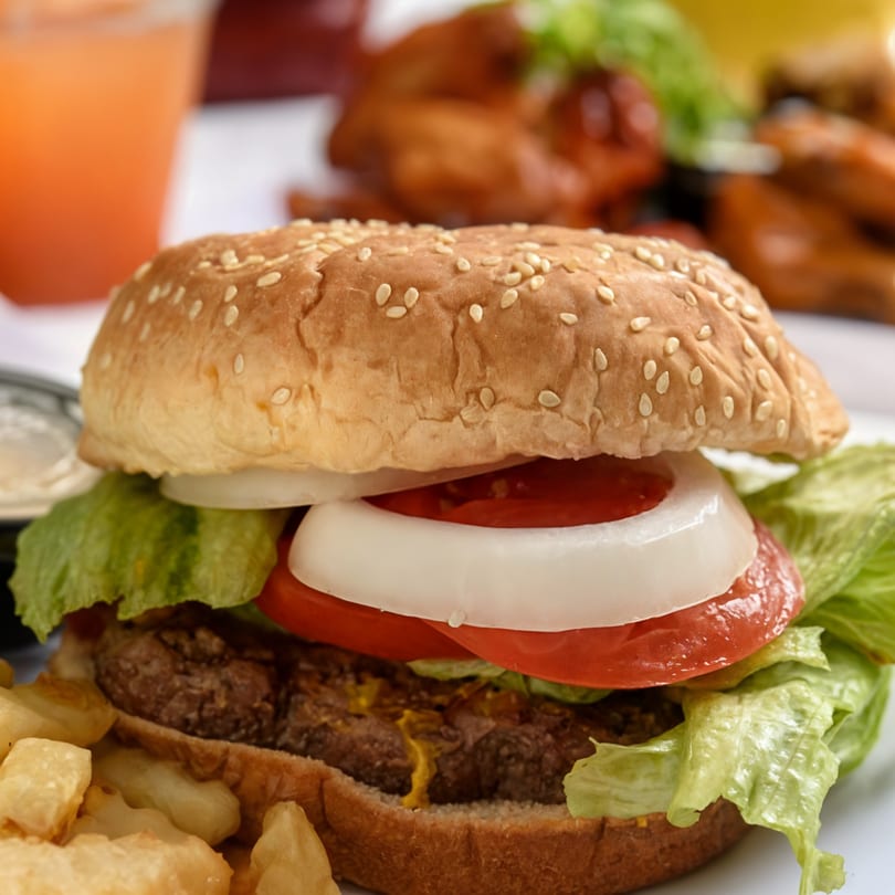 Close-up of a burger with lettuce, tomato, and onion, with fries and a drink.