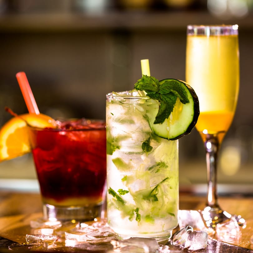 Three colorful cocktails: a red drink with orange, a mojito with cucumber, and a yellow drink.
